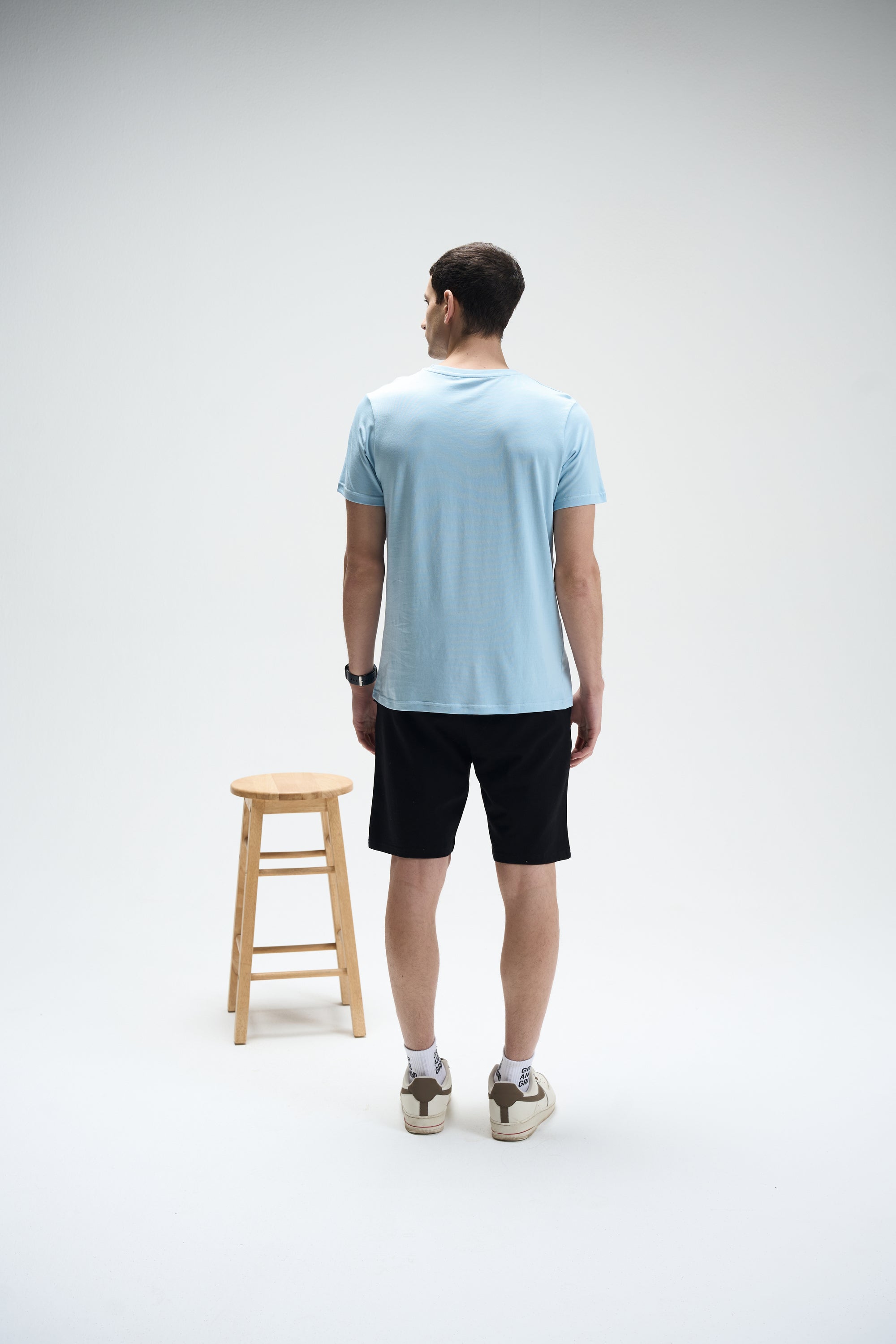 Man wearing powder blue SoftLuxe Crew T-shirt and black shorts, standing next to wooden stool on white background.